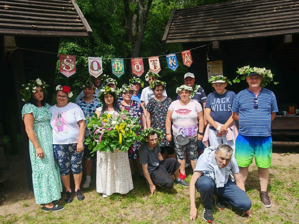 Grupowe zdjęcie uczestników plenerowego spotkania z własnoręcznie wykonanymi wiankami pod banerem „Sobótka”.