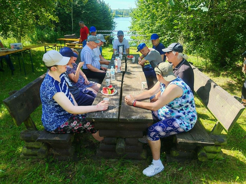 Uczestnicy pikniku siedzą przy drewnianym stole i wspólnie jedzą arbuza w tle widać jezioro.