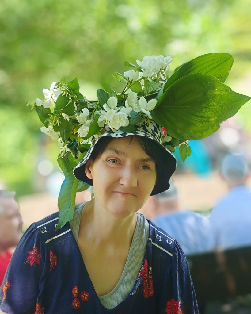 Uczestniczka prezentuje kapelusz ozdobiony wiankiem z białych kwiatów i zielonych liści podczas plenerowego spotkania.