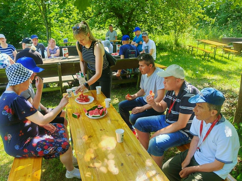 Uczestnicy pikniku siedzą przy drewnianym stole i wspólnie jedzą arbuza podczas spotkania nad jeziorem