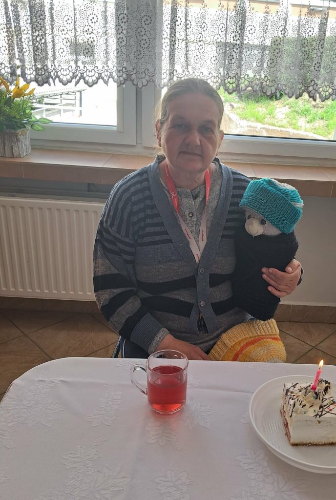 Uczestniczka spotkania urodzinowego siedzi przy stole z kawałkiem tortu i herbatą, trzymając pluszowego misia w czapce."