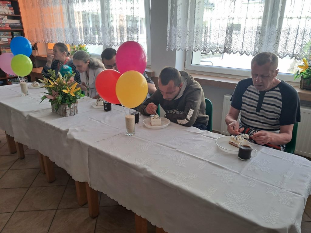 "Uczestnicy spotkania urodzinowego siedzą przy wspólnym stole z tortem, napojami i kolorowymi balonami."