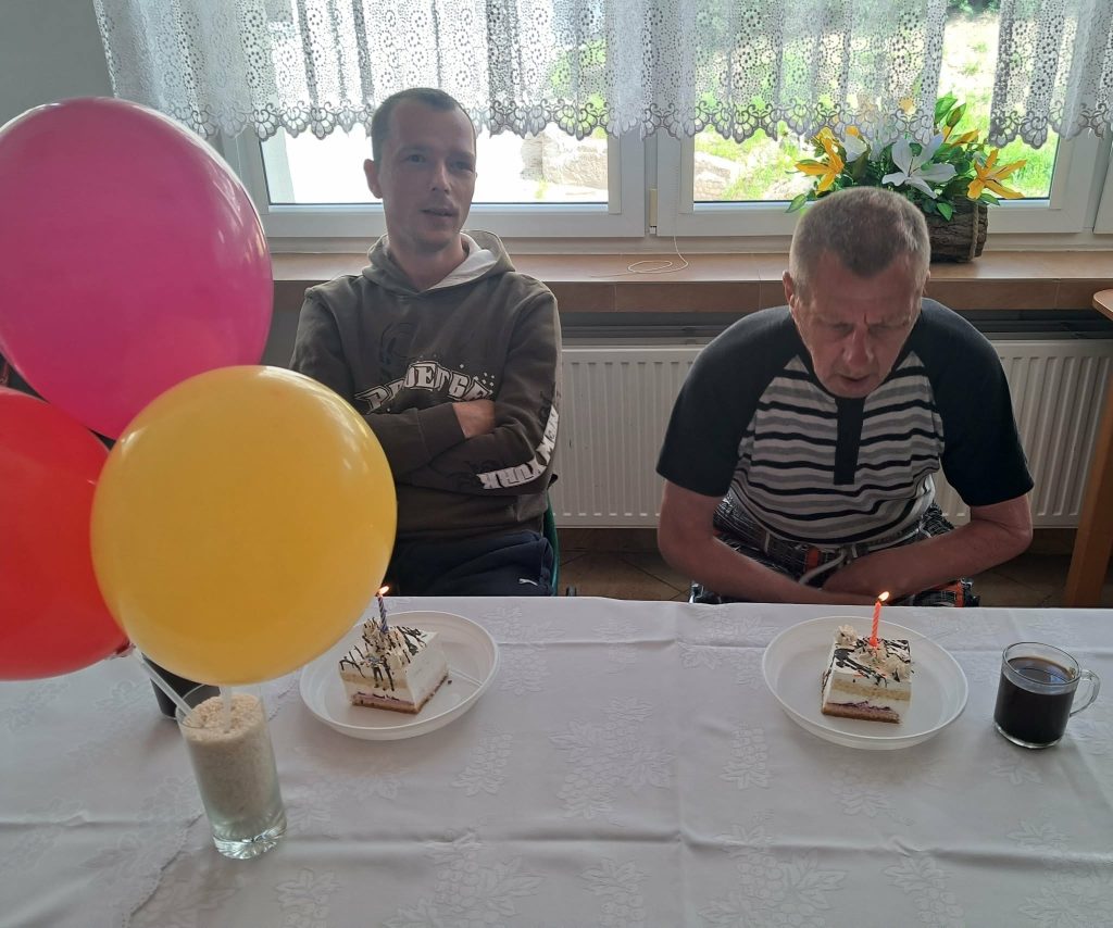 "Dwóch uczestników obchodzi urodziny przy stole z kawałkami tortu ze świeczkami oraz kolorowymi balonami."