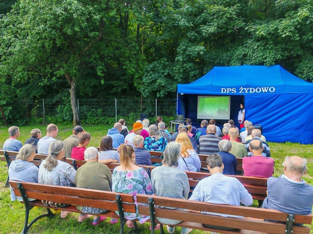 Mieszkańcy DPS Żydowo oglądają film podczas kina plenerowego na świeżym powietrzu; seans odbywa się pod niebieskim namiotem z ekranem.