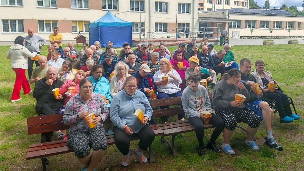Mieszkańcy DPS Żydowo siedzą na ławkach przed budynkiem ośrodka i uczestniczą w kinie plenerowym, trzymając popcorn oraz napoje.