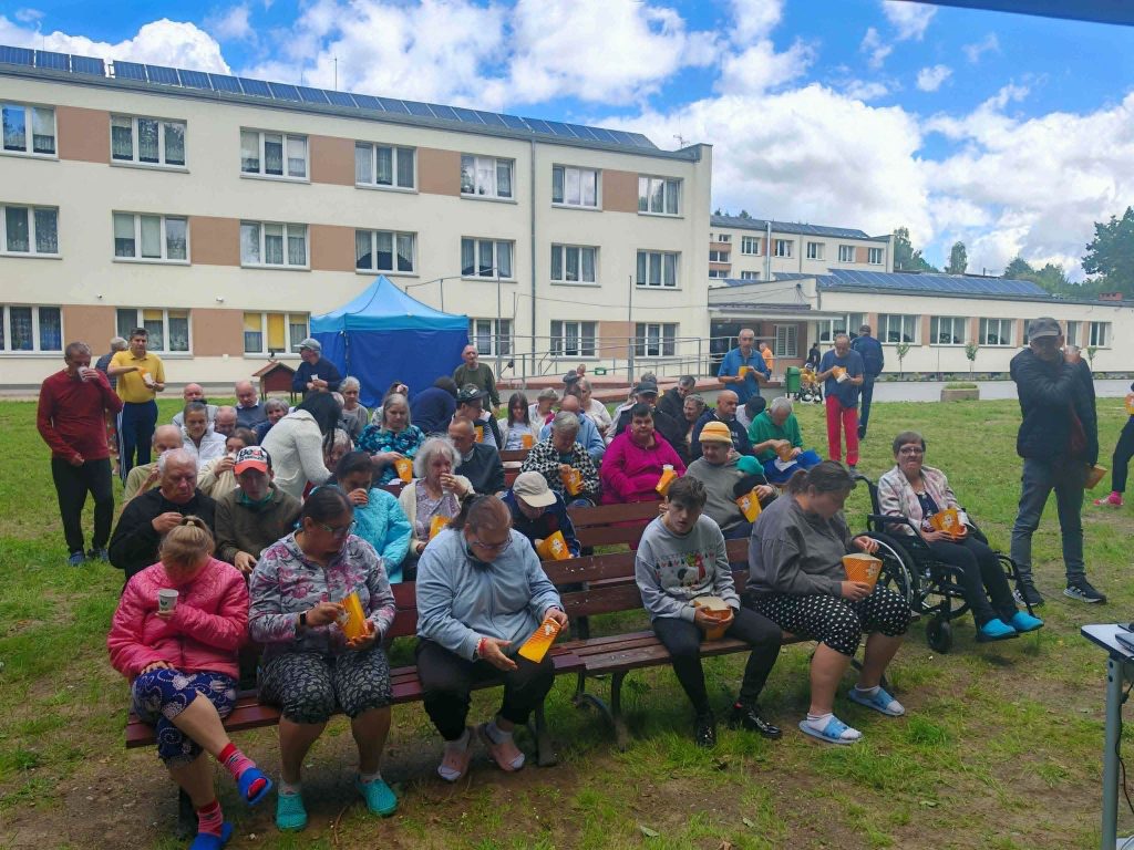 . Uczestnicy kina plenerowego w DPS Żydowo zajmują miejsca na ławkach przed rozpoczęciem seansu, trzymając popcorn i napoje.