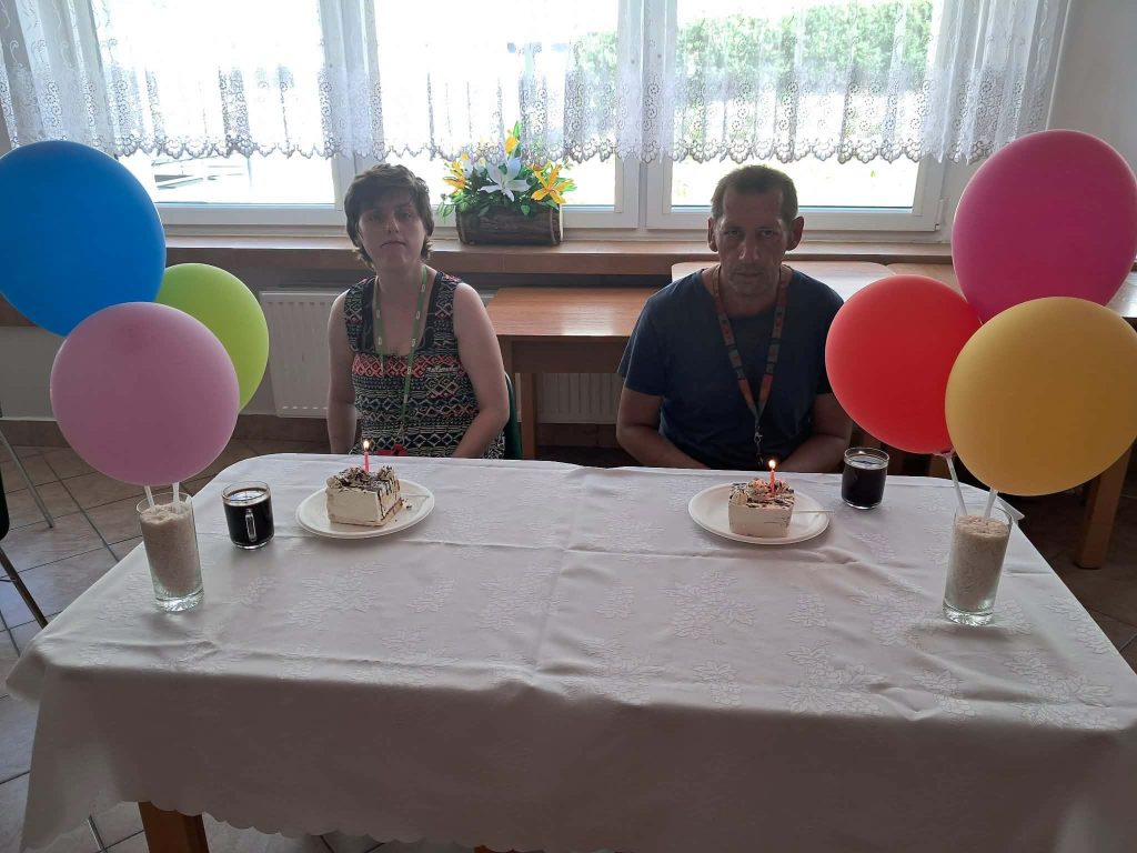 Dwoje mieszkańców DPS siedzi przy udekorowanym stole z tortami urodzinowymi i zapalonymi świeczkami; po bokach stołu stoją kolorowe balony.