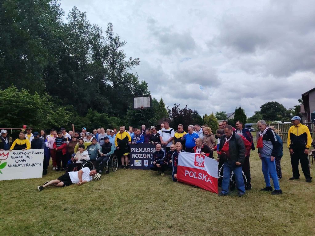 Grupowe zdjęcie uczestników turnieju piłki nożnej w Krągu na boisku; część osób trzyma flagę Polski i tablicę wydarzenia.