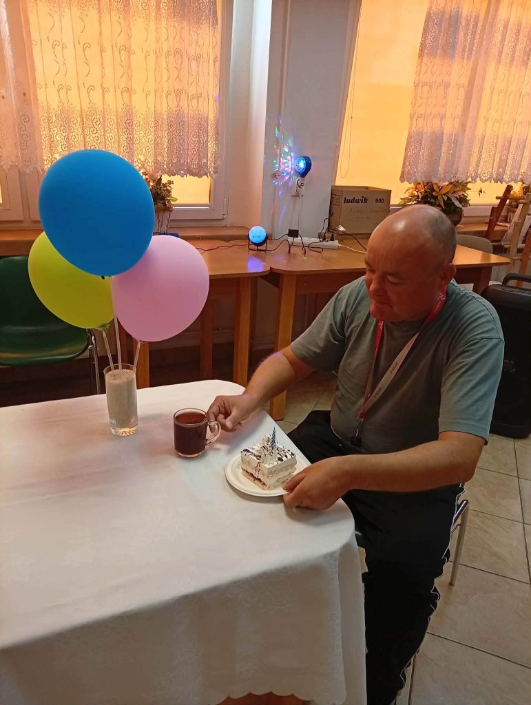 Mieszkaniec Domu Pomocy Społecznej siedzi przy udekorowanym stole podczas urodzinowego poczęstunku. Przed nim znajduje się kawa i kawałek tortu, a na stole stoją kolorowe balony. W tle widać salę oraz dekoracyjne oświetlenie.