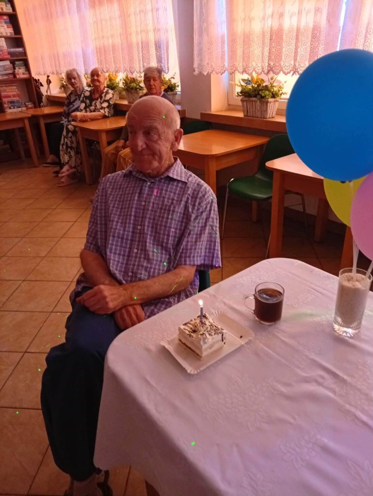Mieszkaniec Domu Pomocy Społecznej siedzi przy stole z urodzinowym ciastkiem ze świeczką i filiżanką kawy. Obok stoją kolorowe balony, a w tle pozostali mieszkańcy uczestniczą w spotkaniu urodzinowym w świetlicy.