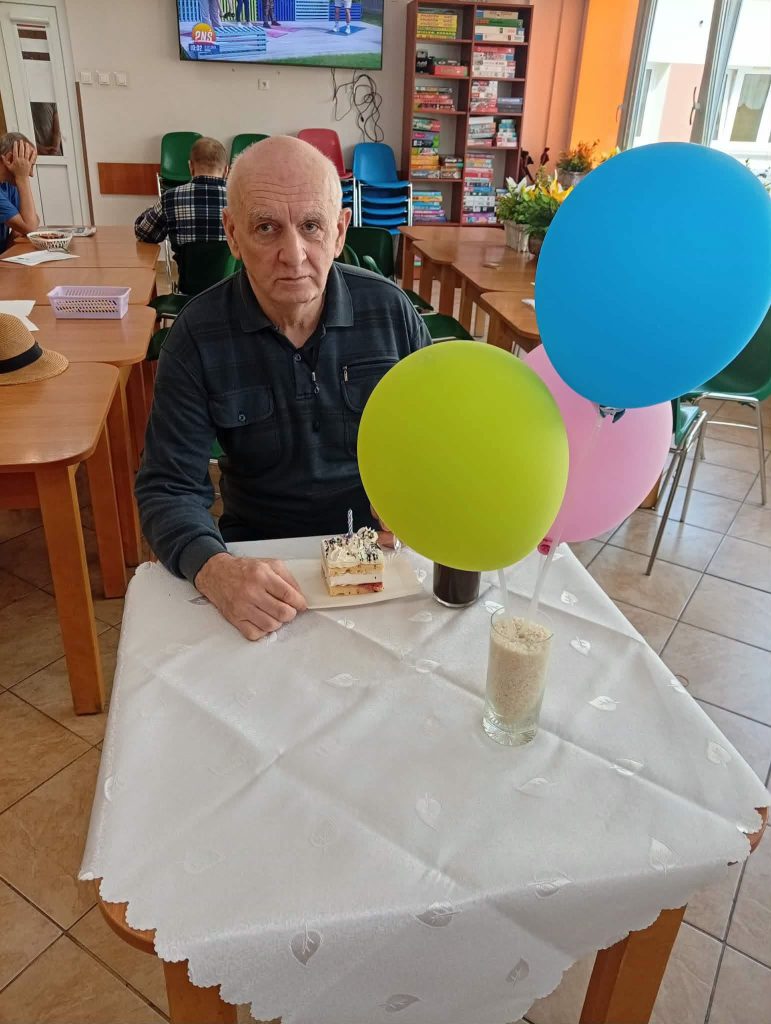 Mieszkaniec DPS siedzi przy stole z urodzinowym ciastkiem i kolorowymi balonami podczas urodzinowego spotkania
