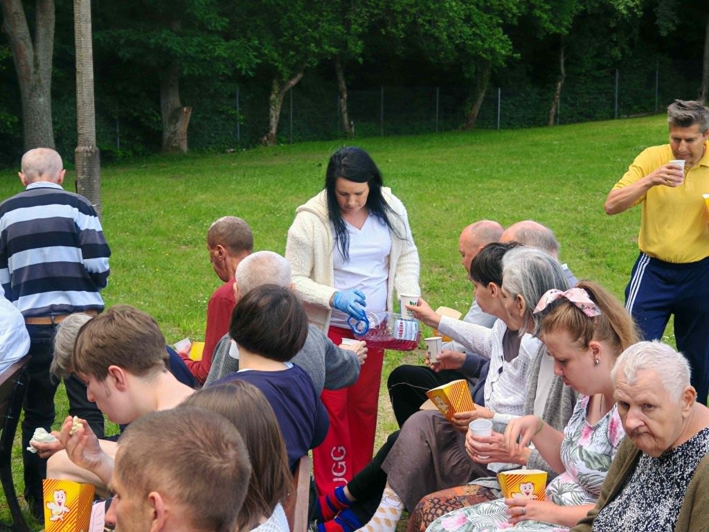 Pracownica DPS Żydowo nalewa mieszkańcom napoje podczas kina plenerowego; uczestnicy siedzą na ławkach z popcornem w rękach.